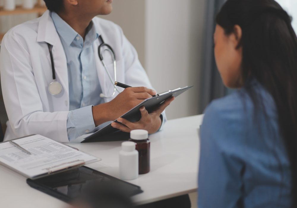Doctor and patient sitting near each other at the wooden desk in clinic. Female physician's pointing to a records form. Medicine concept